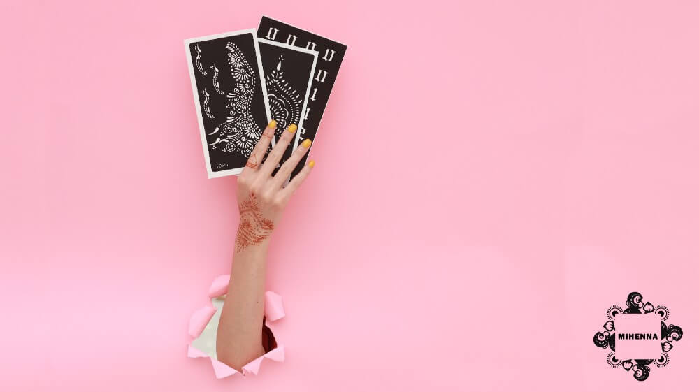Hand breaking through pink paper, holding 3 henna stencils, henna tattoo on back of hand. Mihenna logo in the lower right corner.