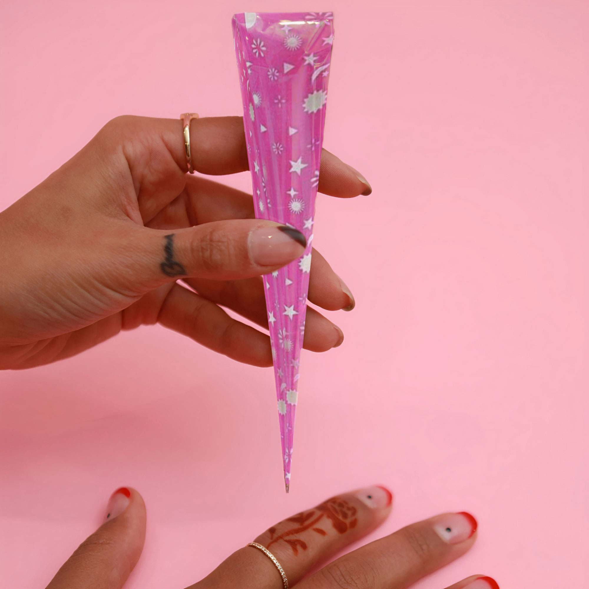 A closeup of a woman’s hands holding a pink henna cone with patterns of stars and moons.