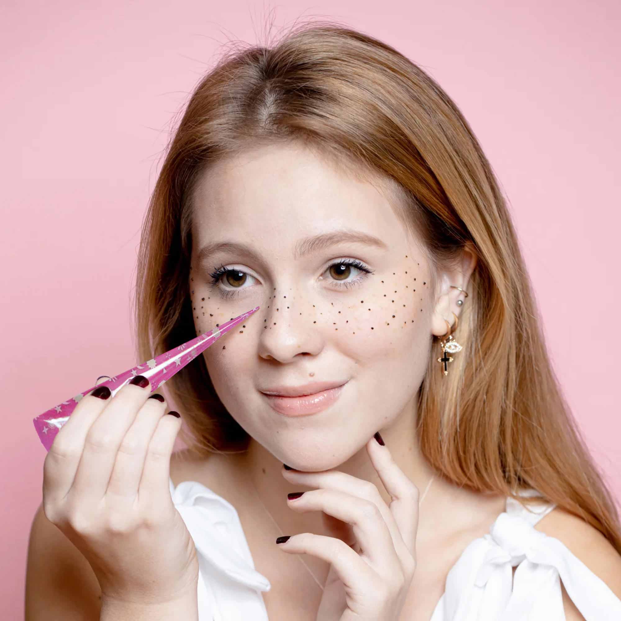 A woman uses a pink henna cone to apply small dots across her nose and cheeks, creating a faux freckle effect.