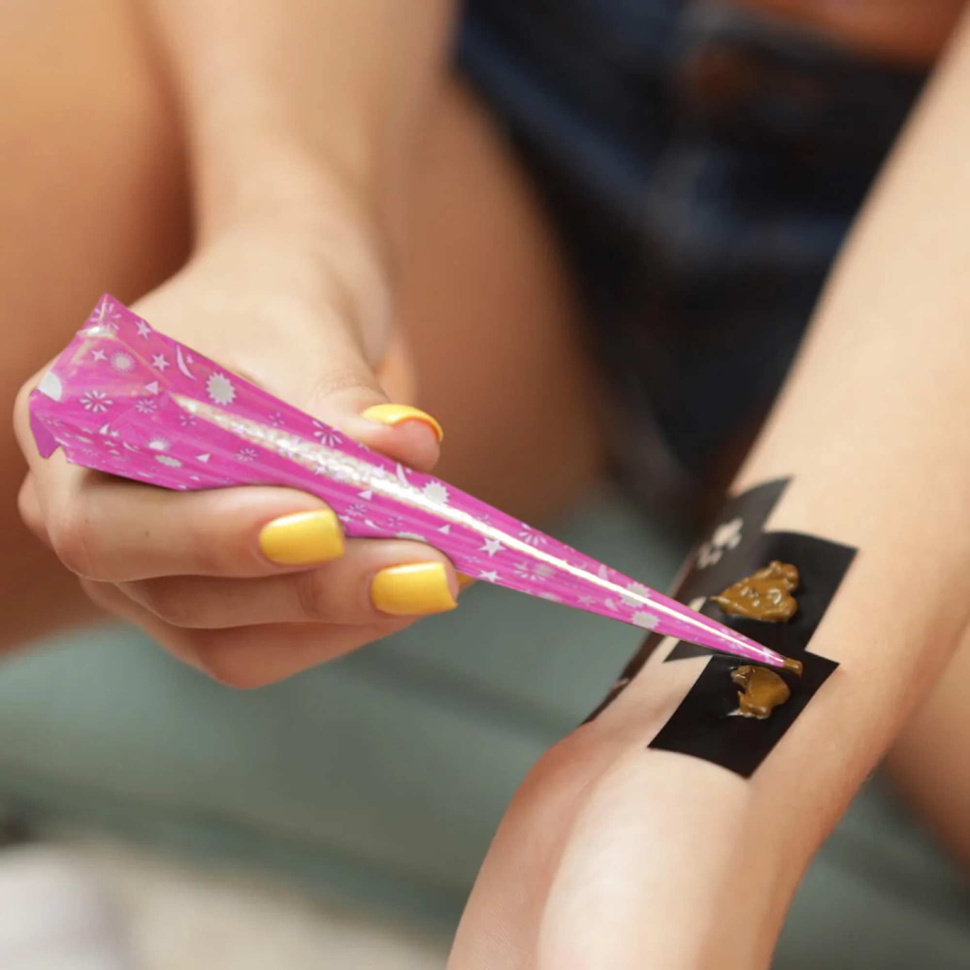 A close-up of a girl’s hand holding a pink, star-patterned henna cone and applying henna paste on her other wrist using stencils.