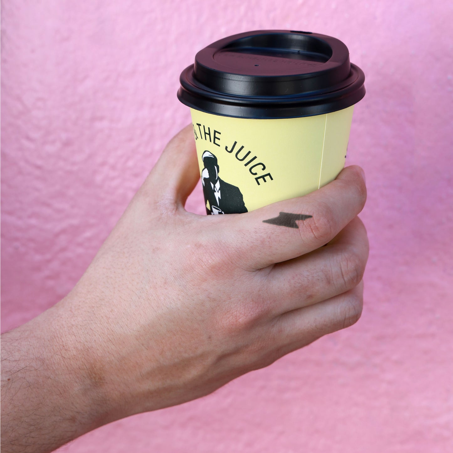 A hand with a thunder tattoo holding a yellow cup with a black closed lid.