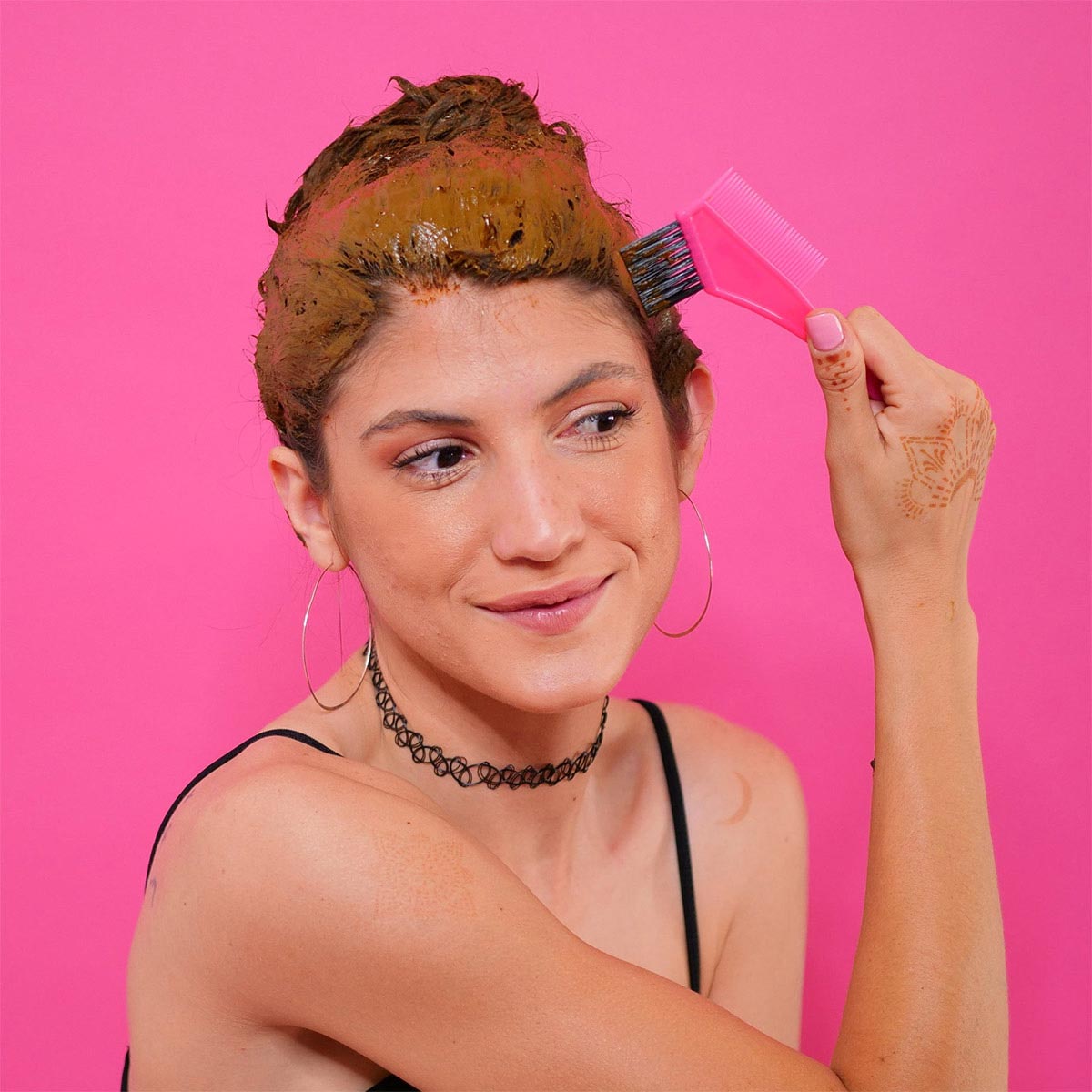 A Woman With Henna Designs on Her Hand Is Applying Henna Paste to Her hair With a Pink Comb Against a Pink Background.